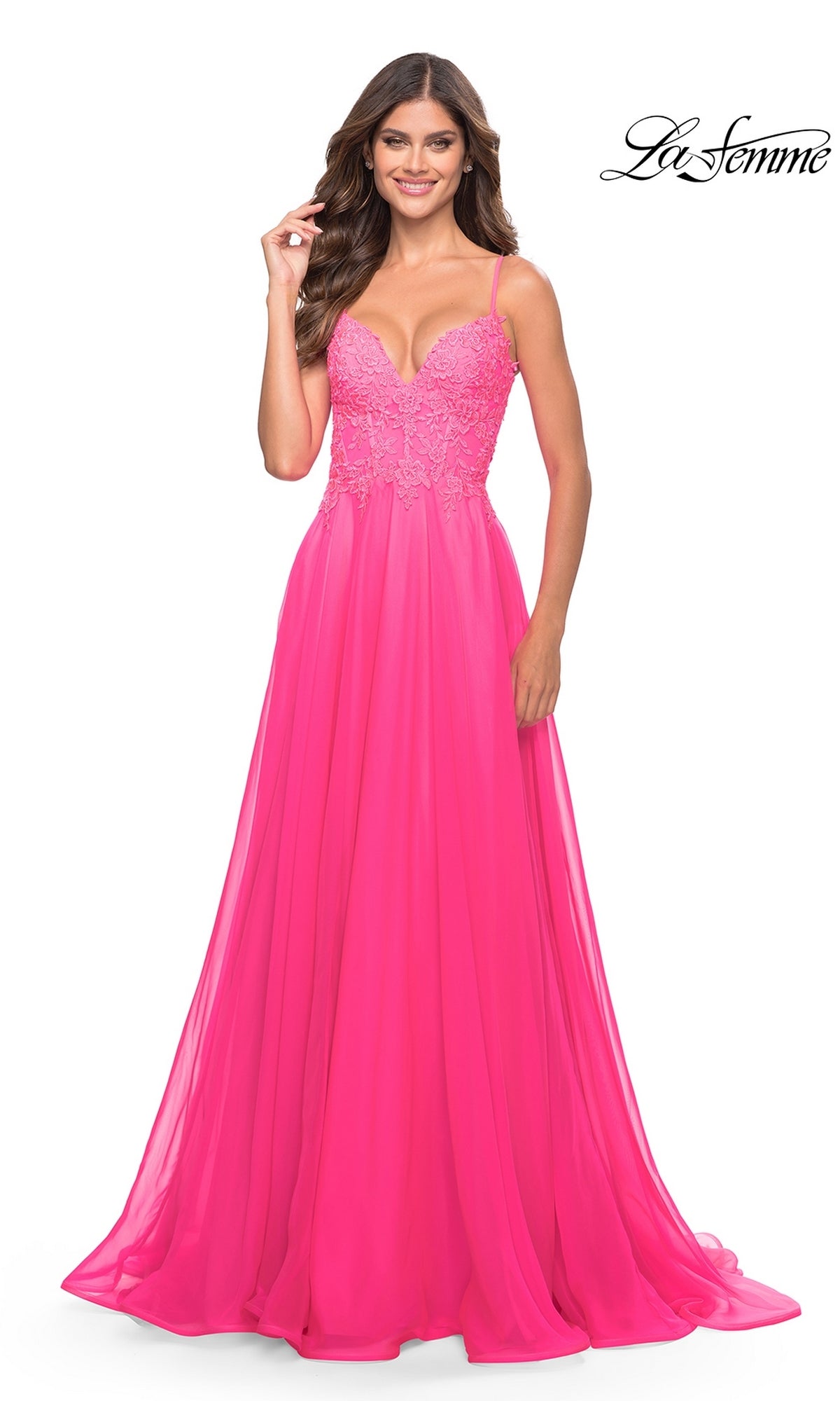 A woman stands smiling in the La Femme Neon Pink Long A-Line Prom Dress 31506, featuring spaghetti straps and a delicately embroidered v-neck bodice on a floor-length formal gown.